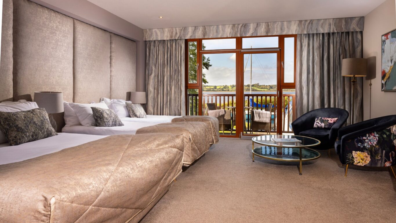 Elegant hotel room with plush beds, floral chairs, cosy lighting, and a scenic balcony view of peaceful waters and greenery.