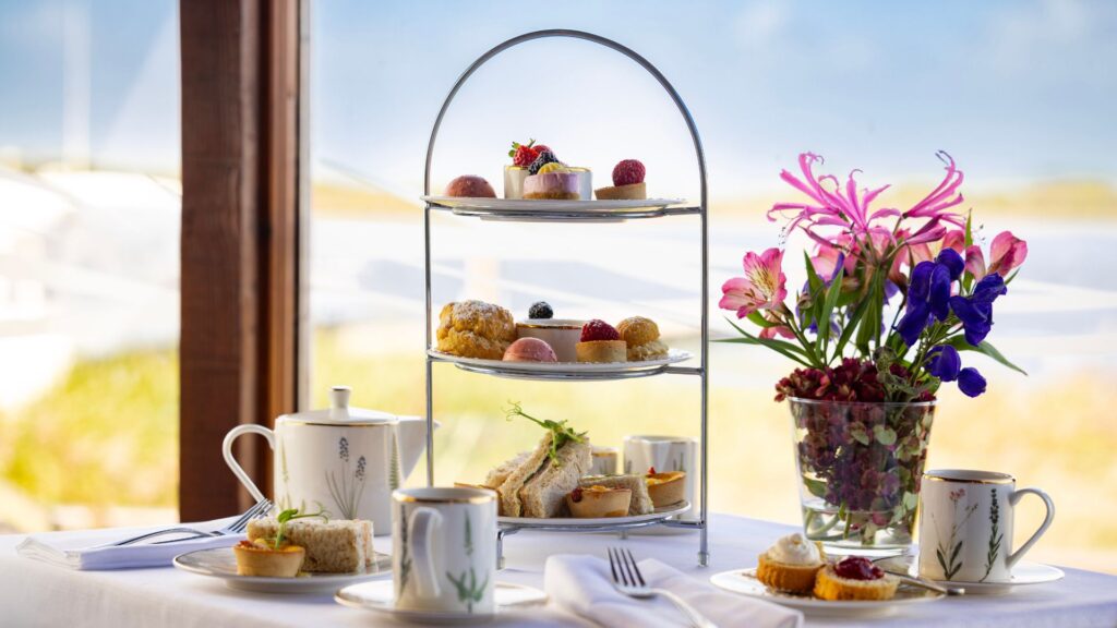Afternoon tea with cakes and sandwiches by a windowside, featuring lush floral centrepiece and elegant tea set.