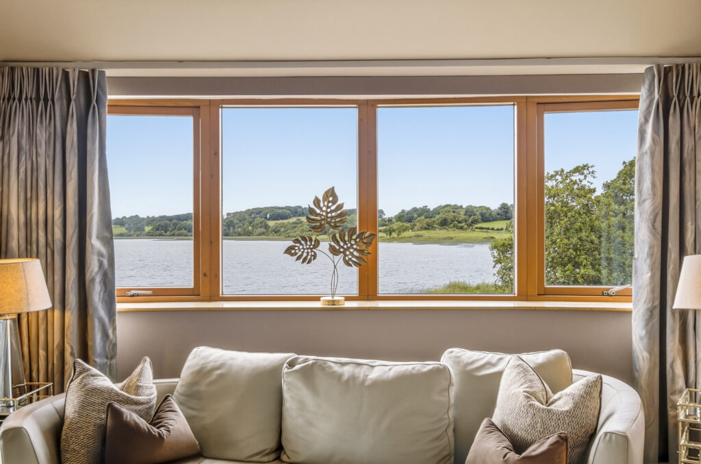 Spacious room with cosy sofa, large window view of serene lake and lush greenery, inviting peaceful retreat.