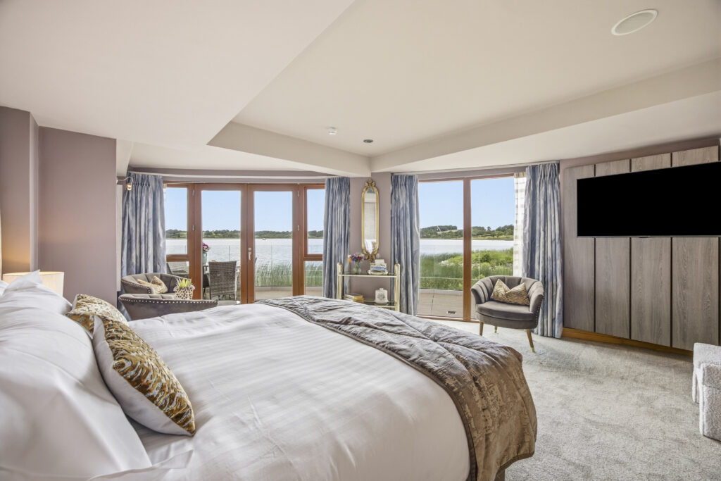 Luxurious hotel room with lake view, plush bedding, stylish armchairs, and large TV, offering serene relaxation.