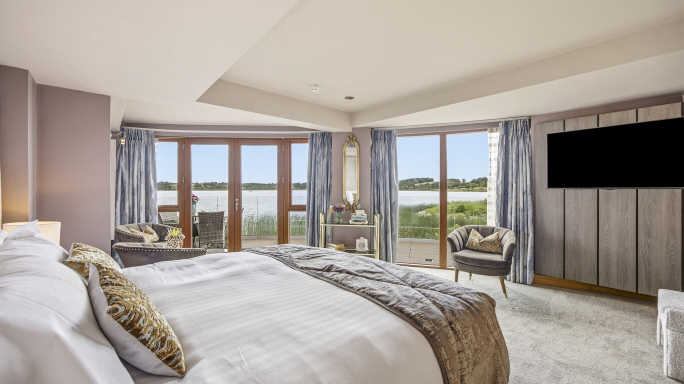Luxurious hotel room with lake view, plush bedding, stylish armchairs, and large TV, offering serene relaxation.