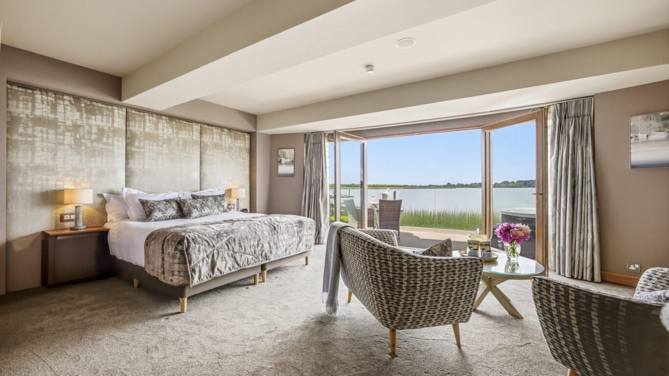 Spacious hotel room with king-sized bed, lake view balcony, comfortable seating, and elegant decor, perfect for relaxation.