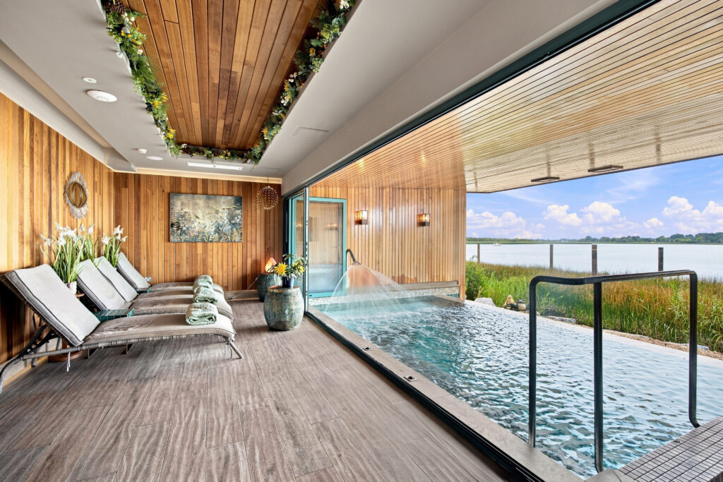 Luxurious spa room with wooden loungers, greenery, indoor pool, and lake view, offering relaxation and tranquility.